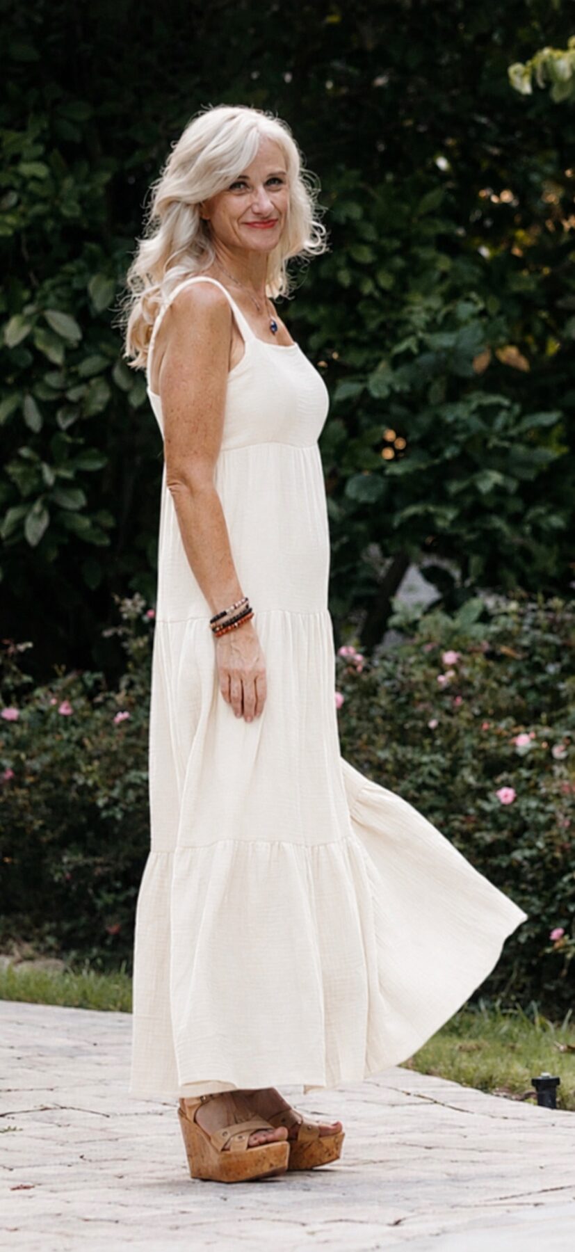 Helen Armstrong in a flowing white maxi dress among garden roses, looking back over her shoulder.